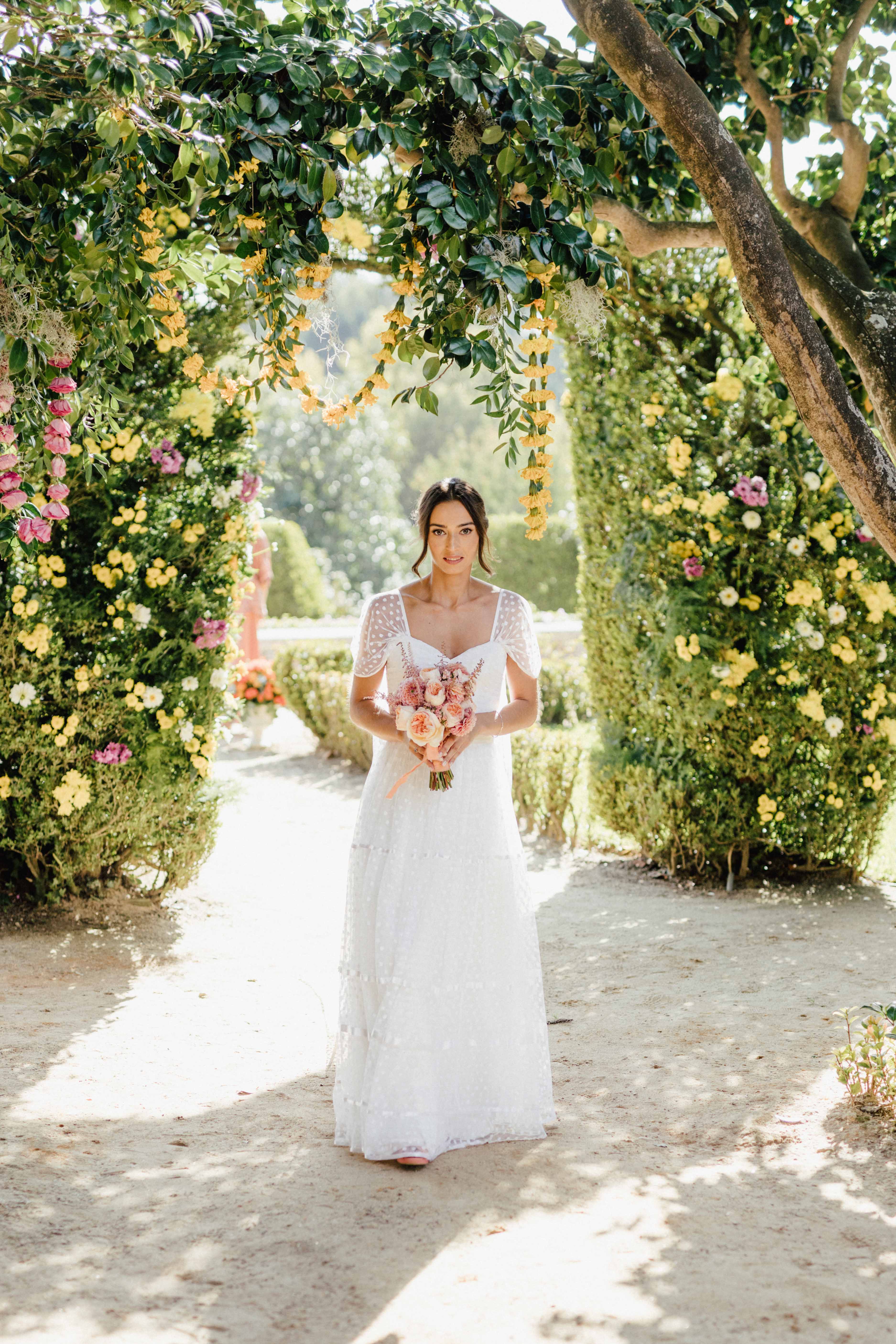 Ana a percorrer o caminho sob arcos de cravo amarelo e cor-de-rosa, vestido branco de polkadot