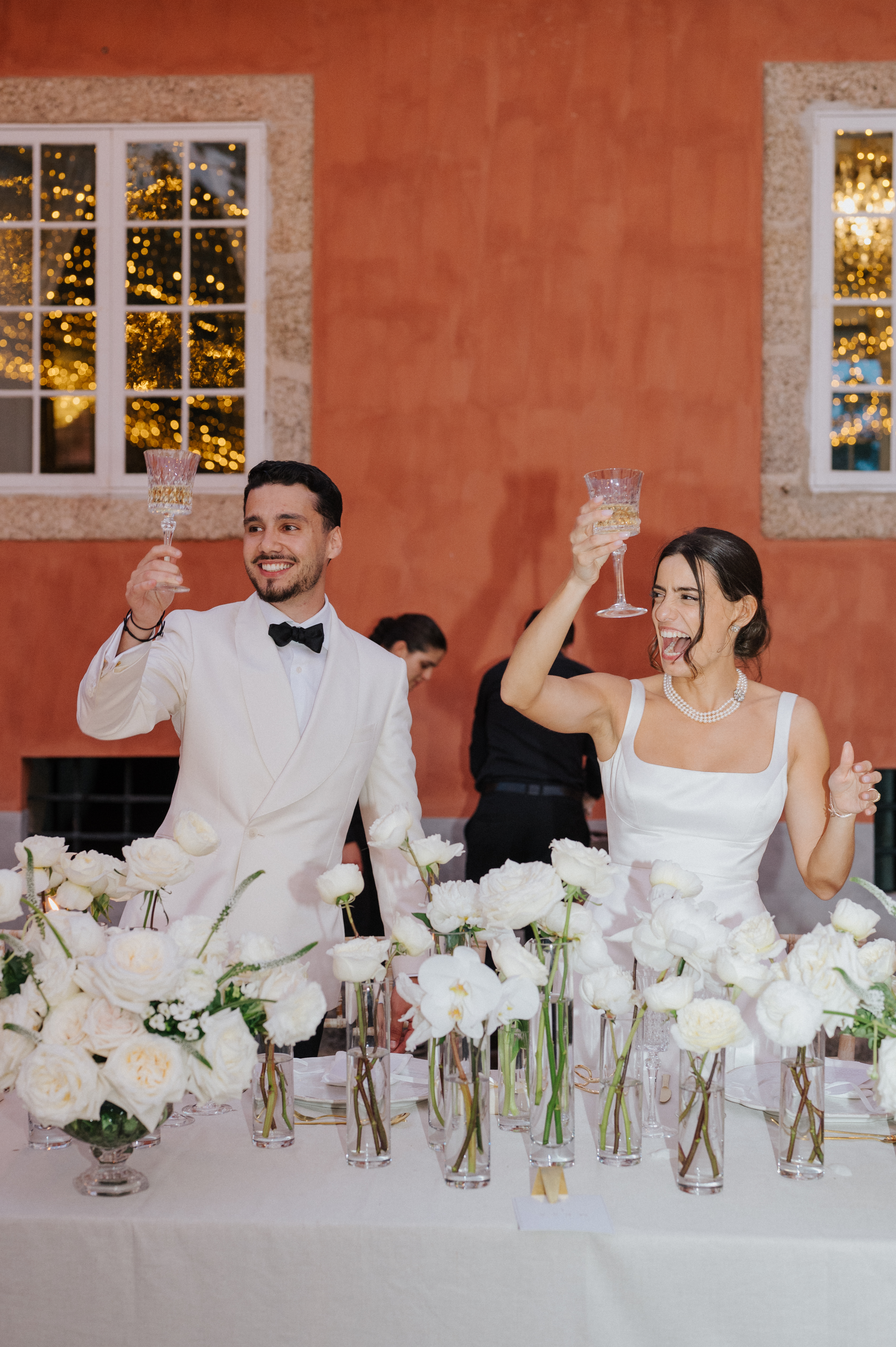 Joana e Francisco a brindar à mesa de honra, vestidos de branco, contra a parede coral de Sezim