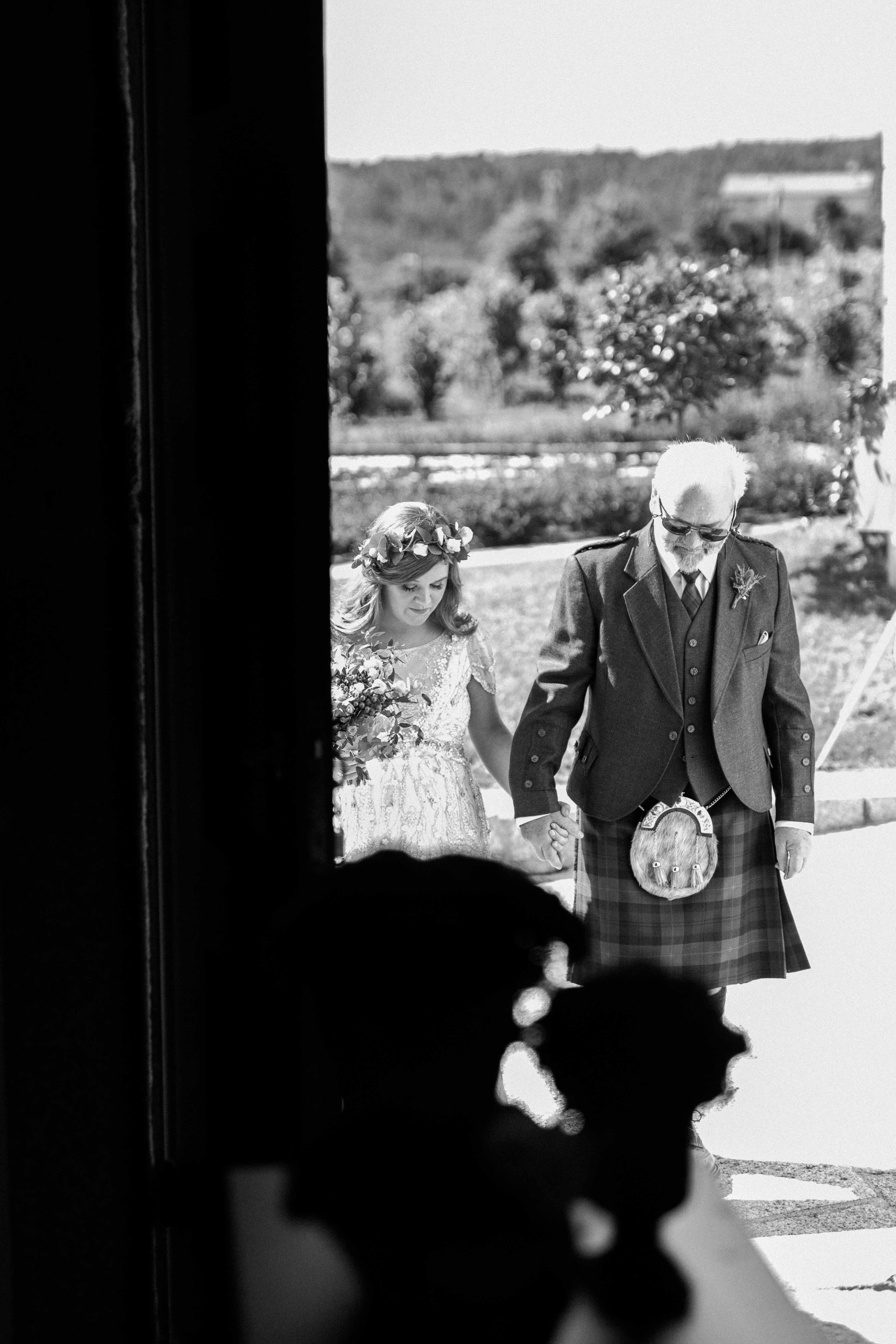 A noiva entra com o pai, em kilt, vista da porta da igreja, em preto e branco
