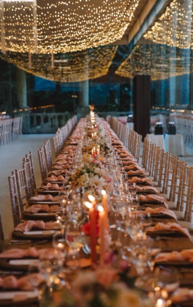 Mesa longa em Colunata, sob um céu de fairy lights, com guardanapos rosa e velas em coral