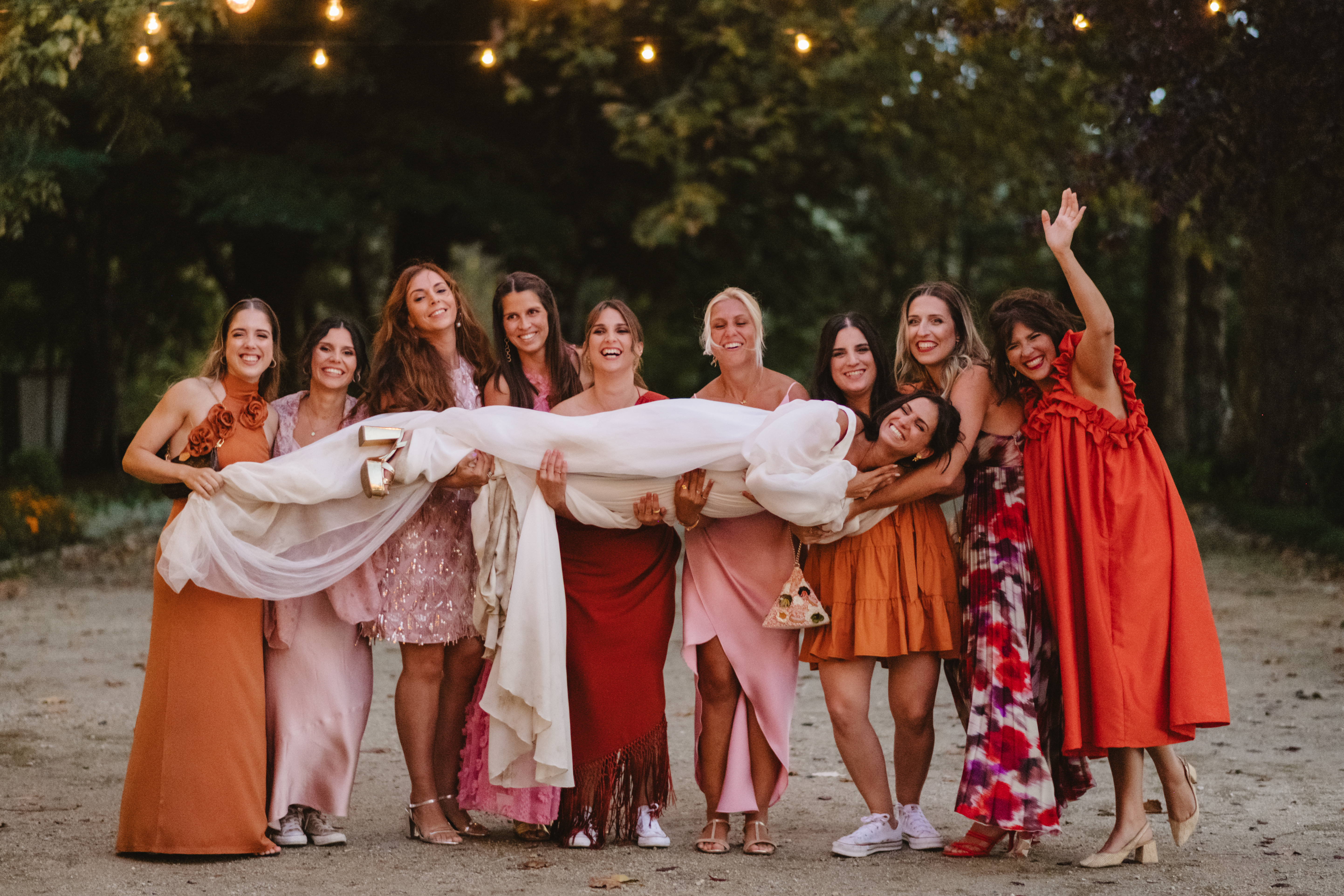 Patrícia levantada do chão por um grupo de amigas em vestidos coral, rosa e laranja, sob fairy lights