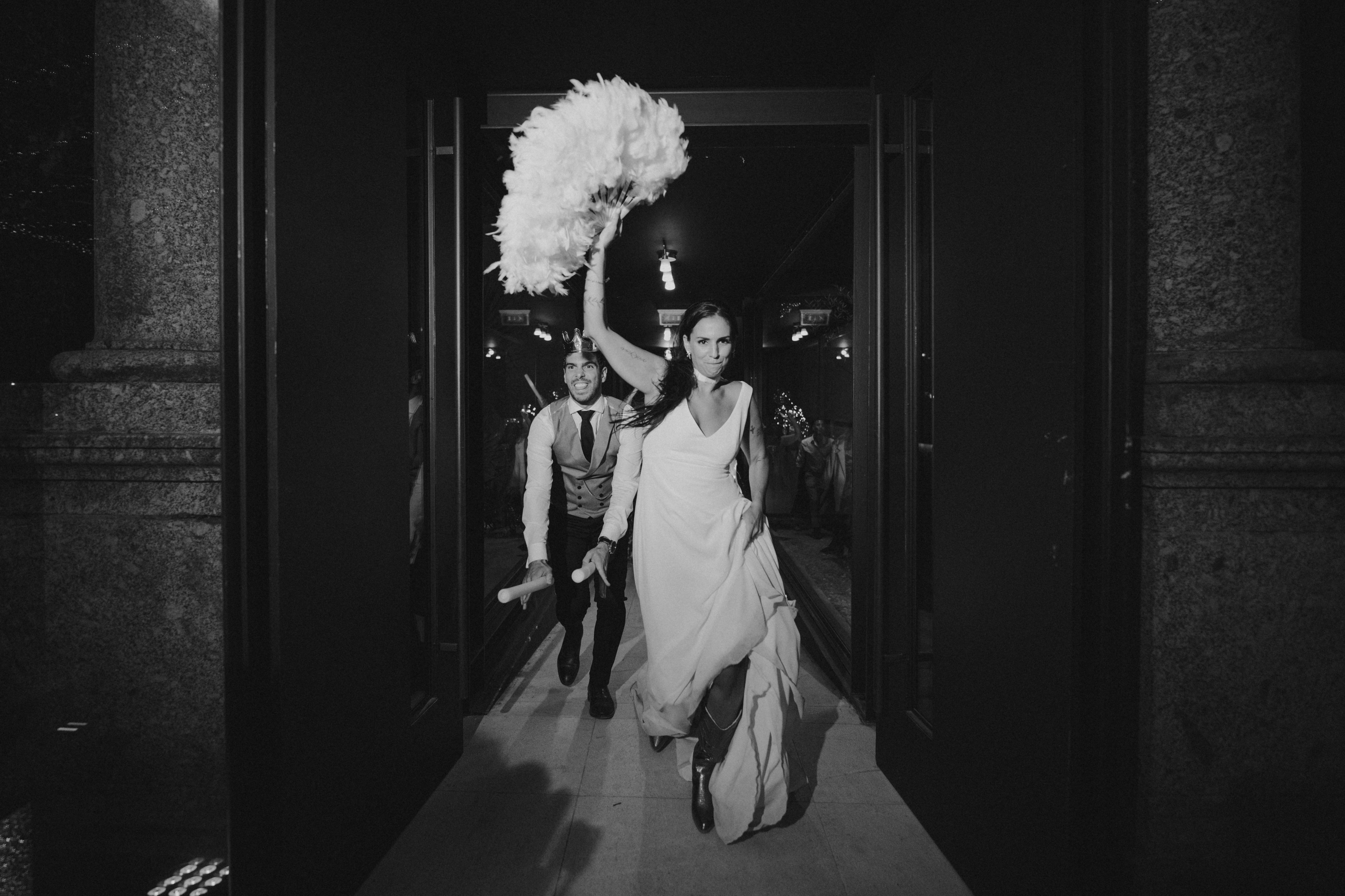 Patrícia e João a sair pela porta principal, ela com um leque de plumas brancas no alto, ele com uma coroa, em preto e branco