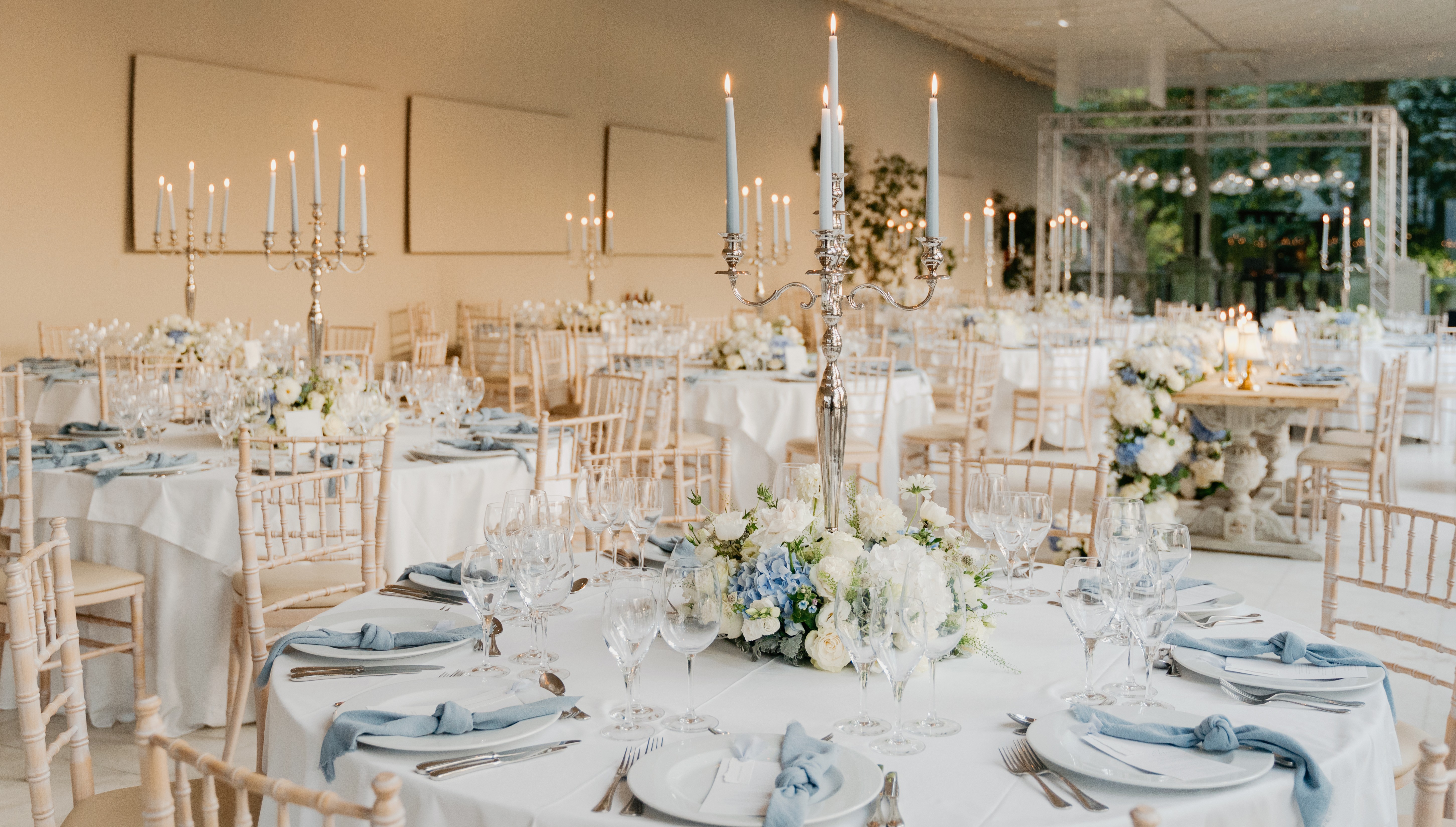 Sala posta para o jantar na Colunata, com mesas redondas em linho branco, candelabros prateados, candeeiros de mesa em latão, e tecto de fairy lights