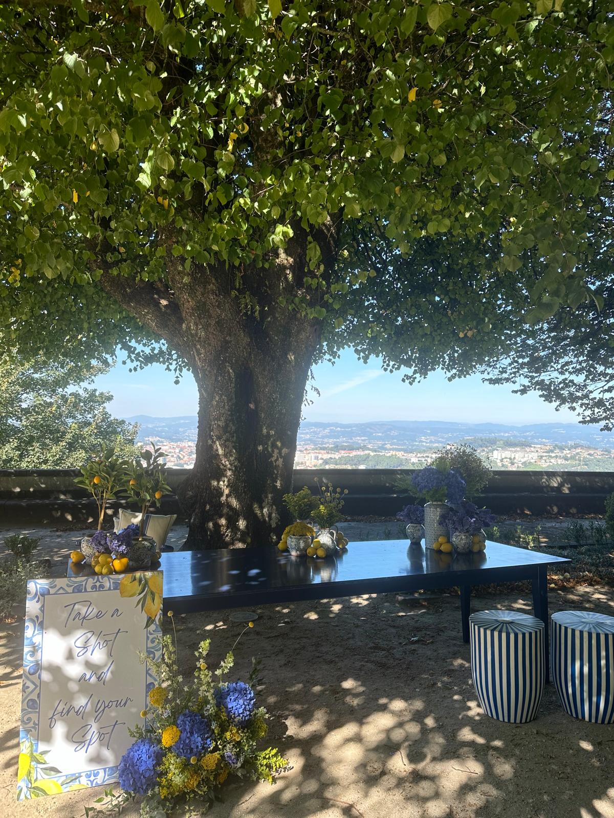 Mesa do cocktail à siciliana, sob uma árvore antiga, com limões em vasos, hortênsias azuis, banquinhos riscados azul e branco, e o cartaz Take a Shot and find your spot, com vista de Braga ao fundo