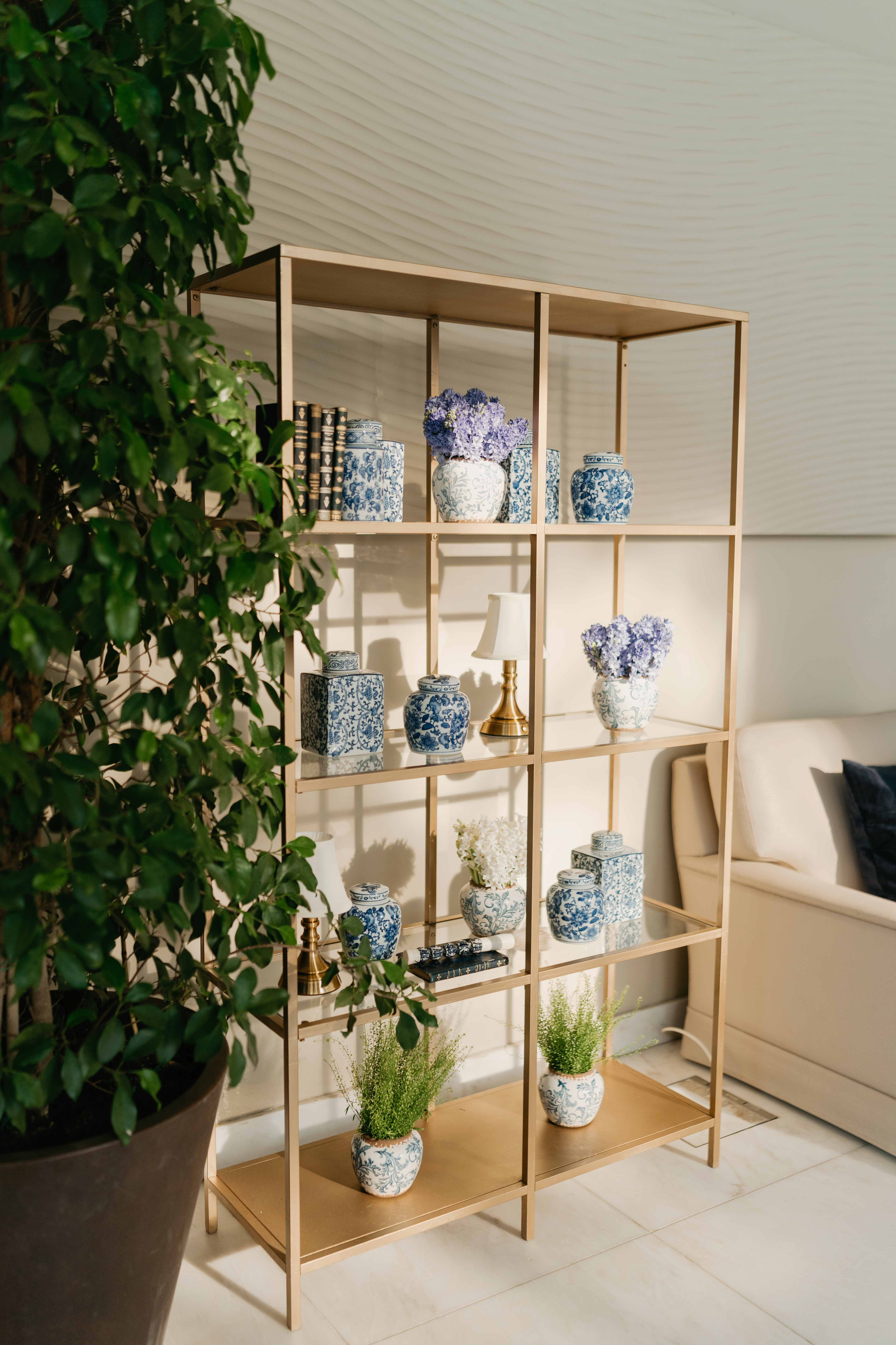 Estante alta em latão com peças de porcelana azul e branca ao estilo chinoiserie, hortênsias roxas, livros antigos, e candeeiros de mesa em latão, num lounge interior