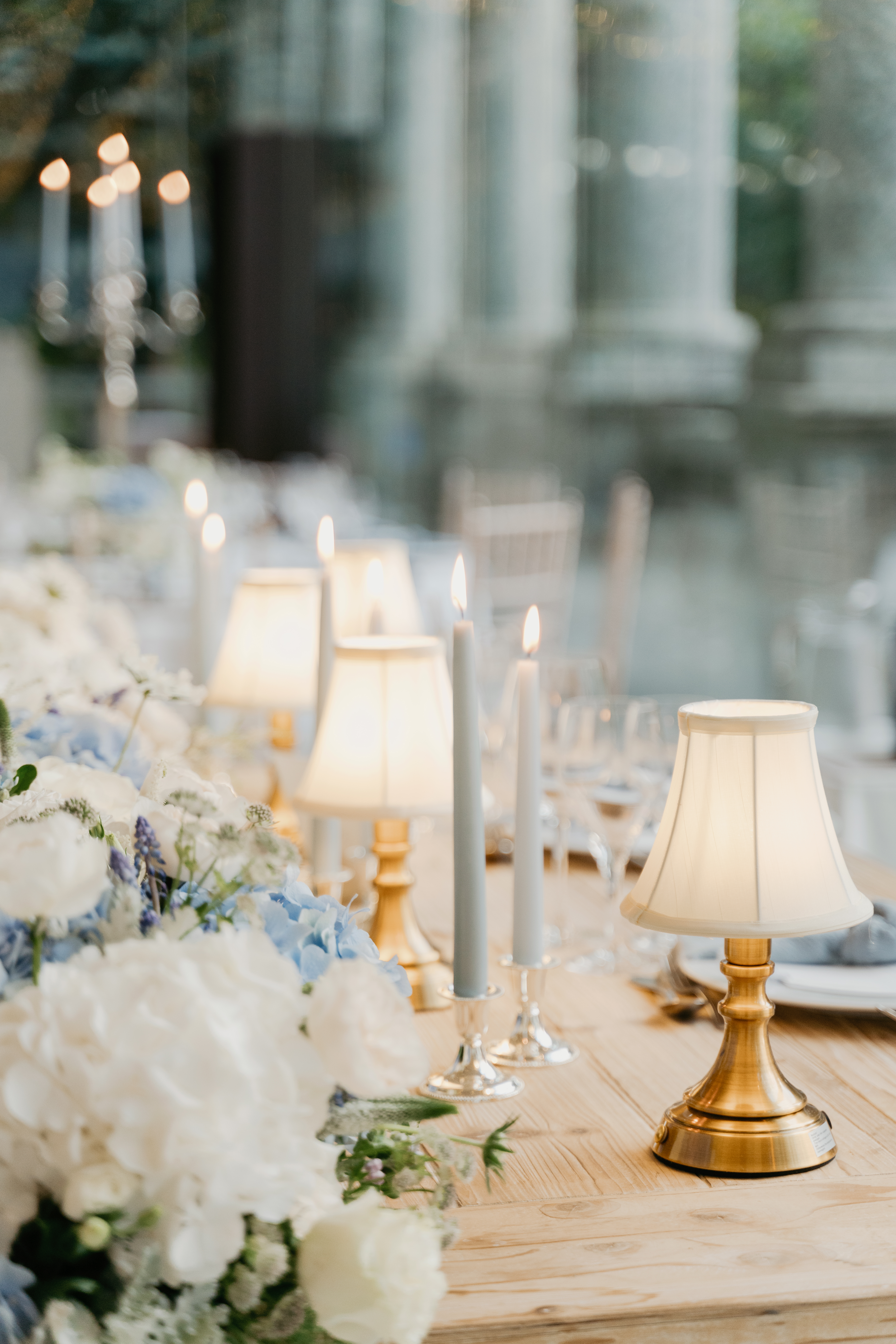 Pormenor da mesa do jantar com candeeiros de mesa em latão de abat-jour creme, velas em tons azuis, hortênsias azuis e rosas brancas, em sequência ao longo de uma mesa em madeira clara