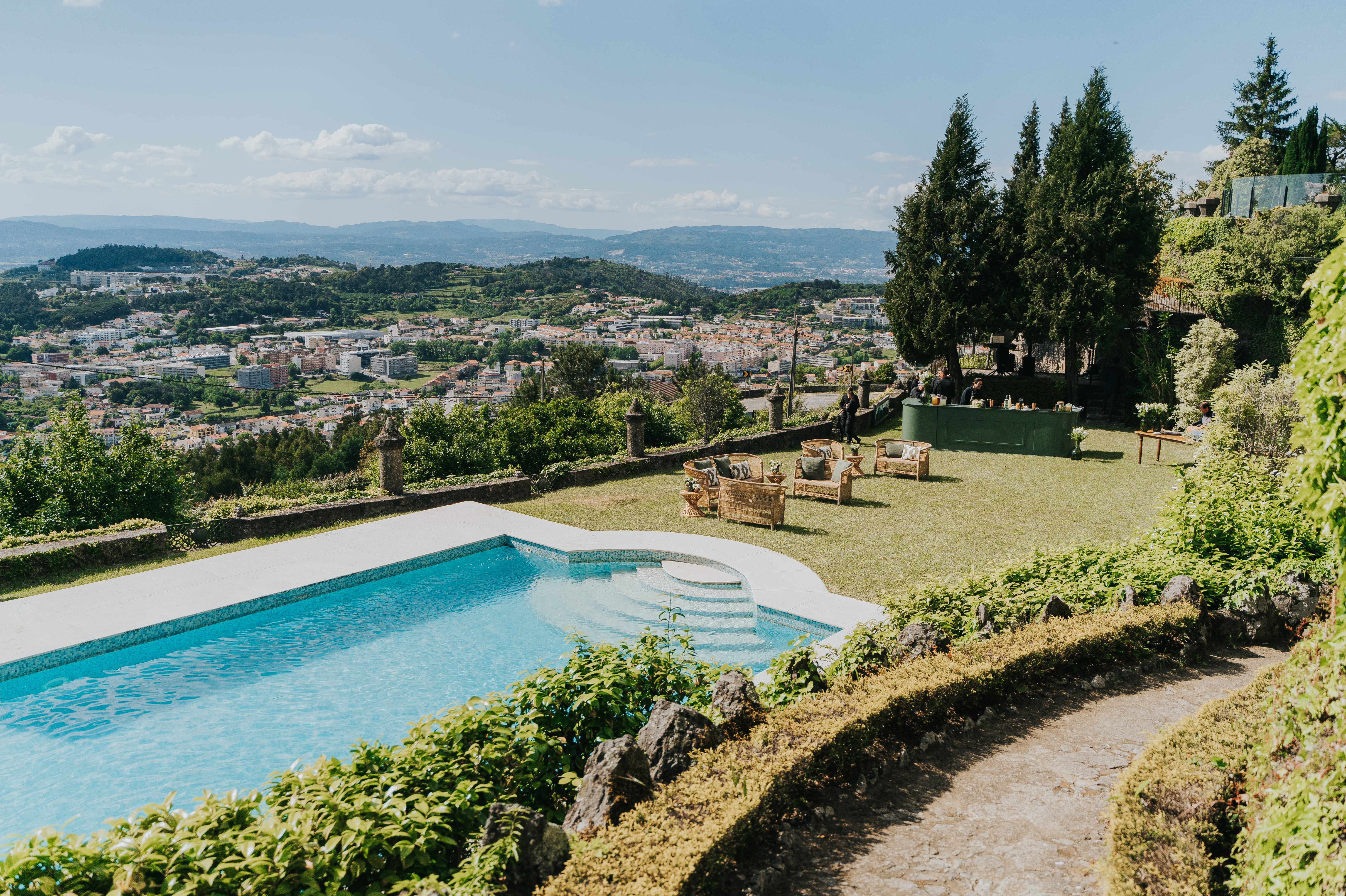Vista aérea da esplanada do Castelo do Bom Jesus, com lounge em rattan, bar verde, piscina, e a paisagem de Braga ao fundo