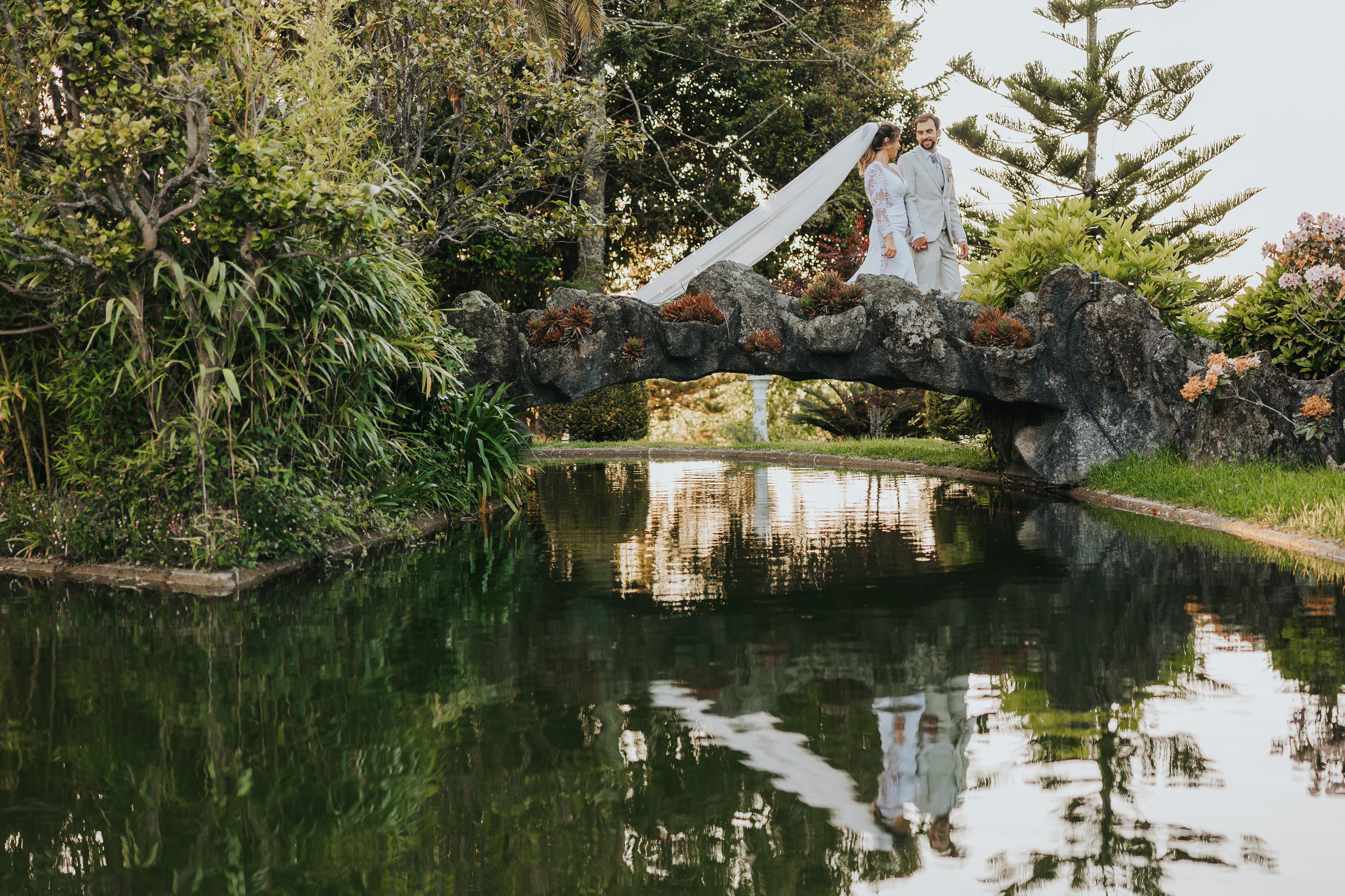 Rita e Hernani na ponte de pedra coberta de suculentas, sobre o lago do jardim do Castelo do Bom Jesus, com reflexo na água
