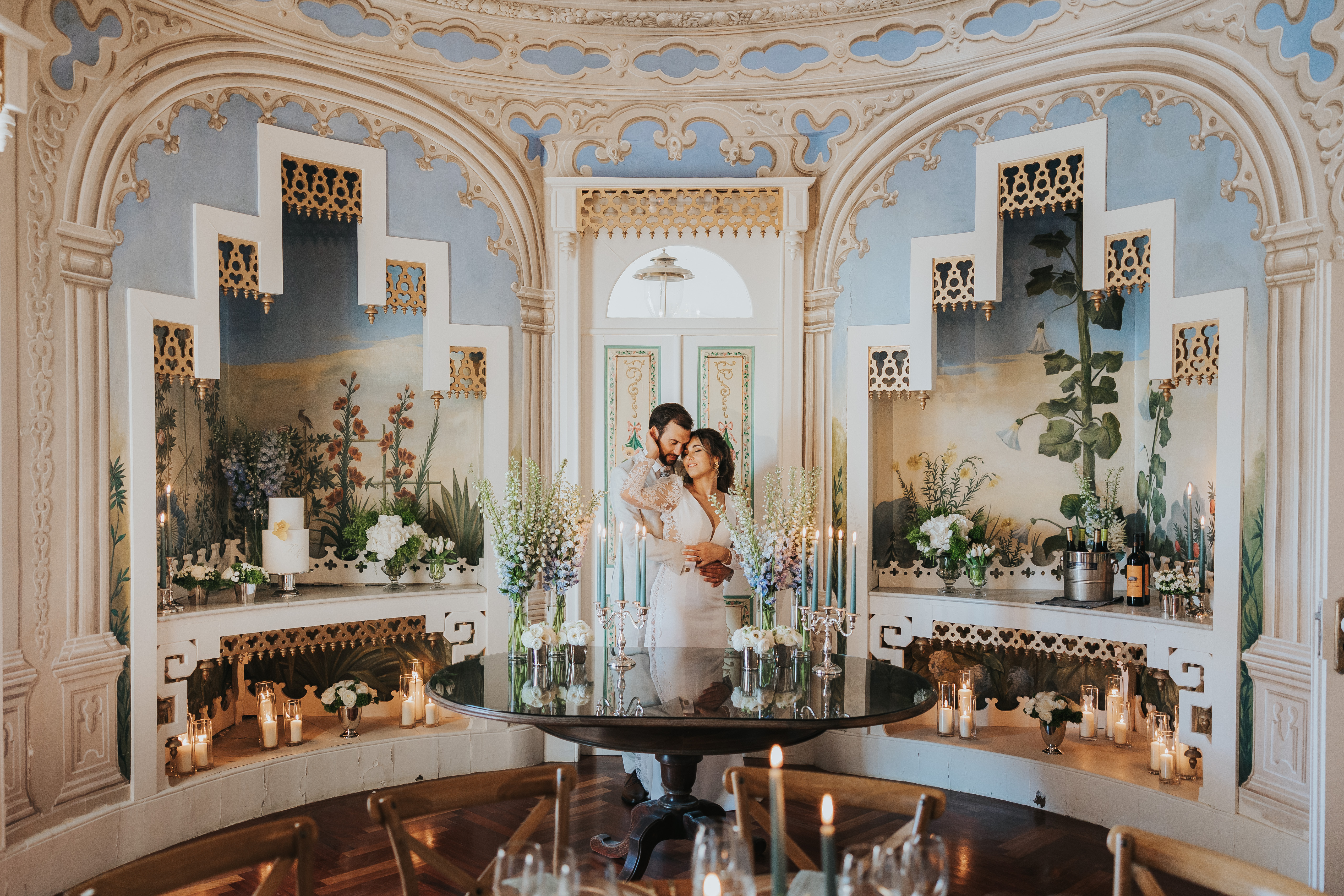 Rita e Hernani num momento íntimo na Sala Árabe, em frente às portas pintadas, com candelabros prateados e arranjos altos de flores brancas e azuis