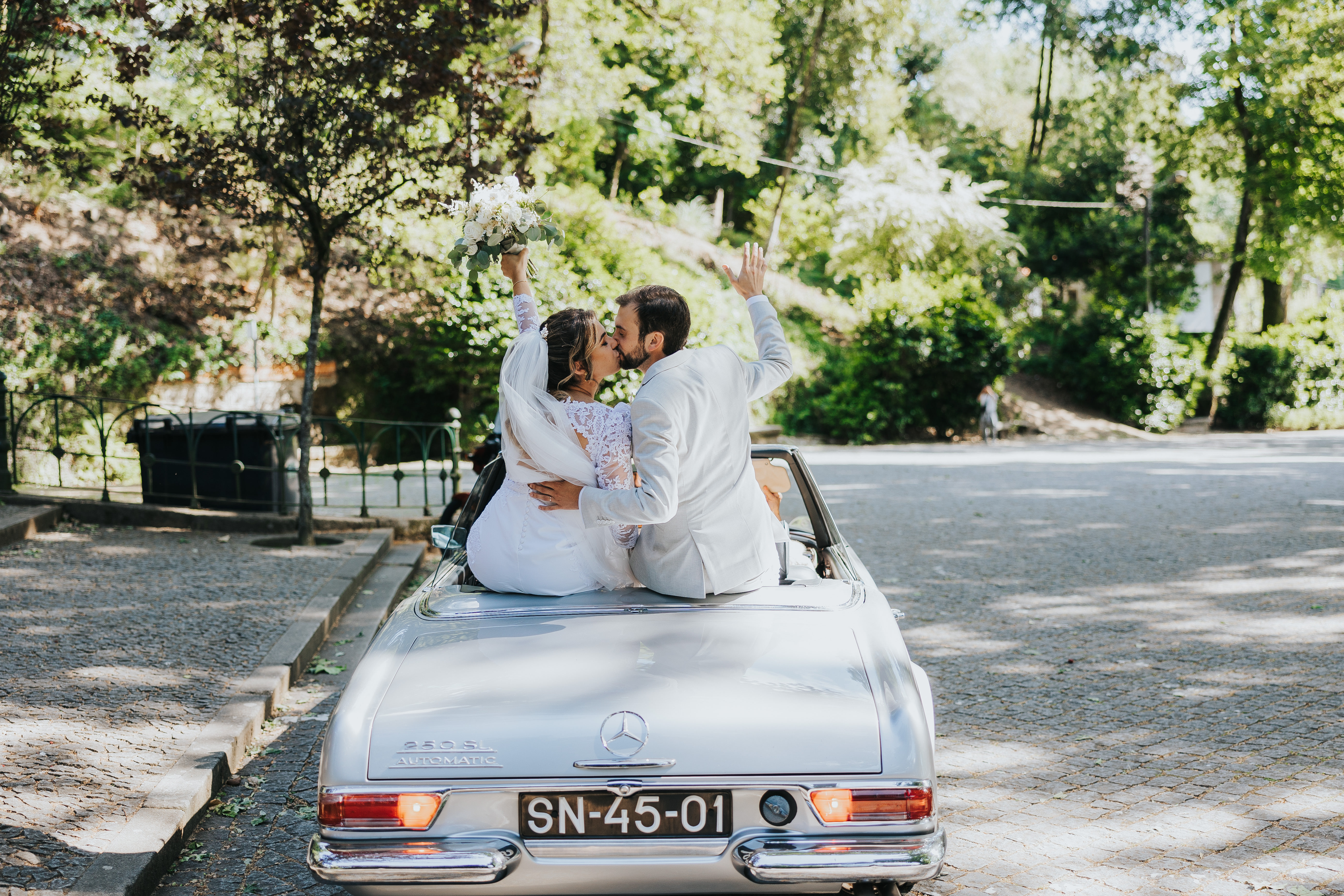 Saída do casal num Mercedes 250 SL descapotável dos anos sessenta, em prata, com a noiva a beijar o noivo e a erguer o ramo de flores brancas